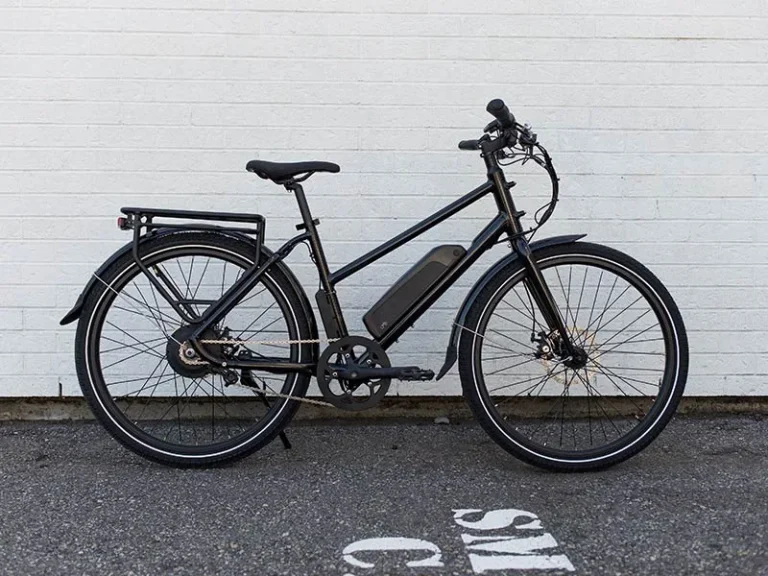 Black electric bicycle against a white brick wall on an asphalt ground. White partial text on ground suggests a parking area.