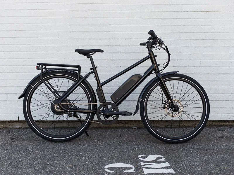 Black electric bicycle against a white brick wall on an asphalt ground. White partial text on ground suggests a parking area.