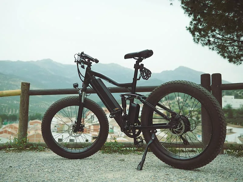 Black mountain bike with thick tires parked on a gravel path, scenic mountain view and overcast sky in the background, peaceful setting.
