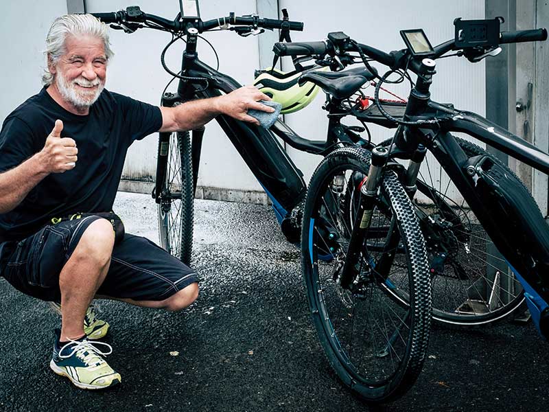 Smiling man kneels, giving a thumbs-up while cleaning bikes with a cloth. He wears a black shirt, shorts, neon sneakers. Gray background.