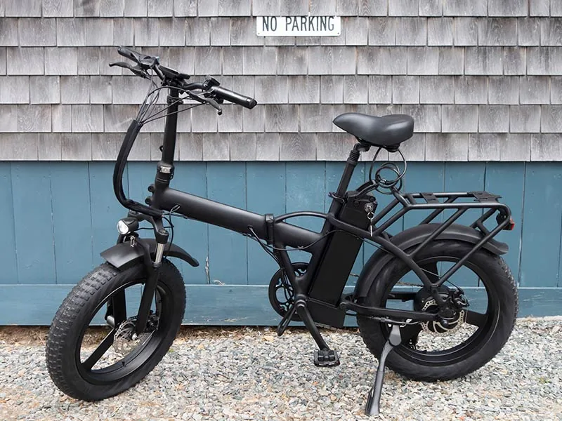 Black electric bike parked on gravel, against a blue and gray shingled wall with a "No Parking" sign.