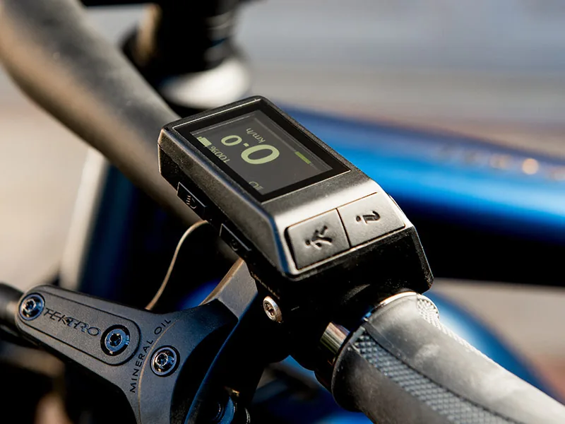Close-up of a bicycle handlebar with a digital display showing 0 km/h. The background is blurred blue and gray, with visible brand text.