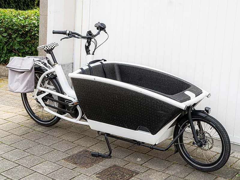 White cargo bike with a large black front compartment on a paved path. Gray panniers on the back and a spotted seat. White panel wall behind.