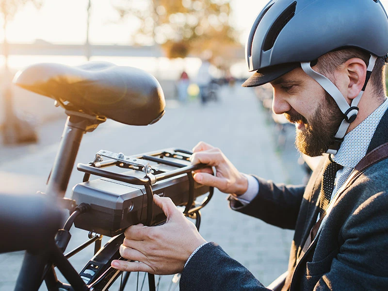 Man with helmet secures battery pack on bike, smiling in warm sunlight. Urban setting with blurred background and cobblestone path.
