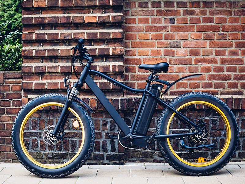 Black electric bike with thick tires and yellow rims, parked against a red brick wall. Urban setting, no visible text or motion.