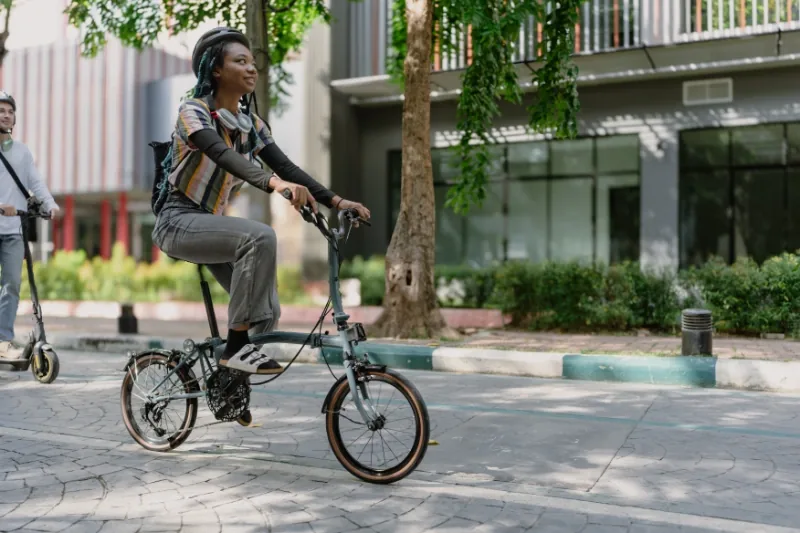 Person with helmet and headphones rides a folding bike in a sunny urban setting, wearing a colorful striped shirt and jeans.