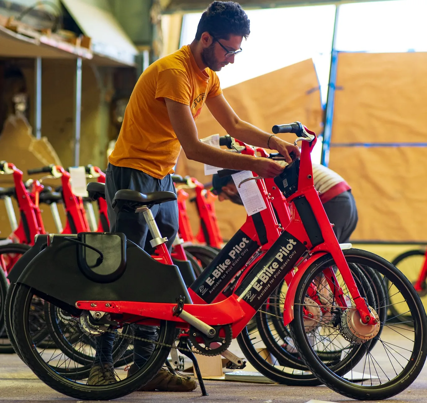 A technician adjusting a red E-Bike Pilot bike in a warehouse, with a row of the same bikes lined up behind him