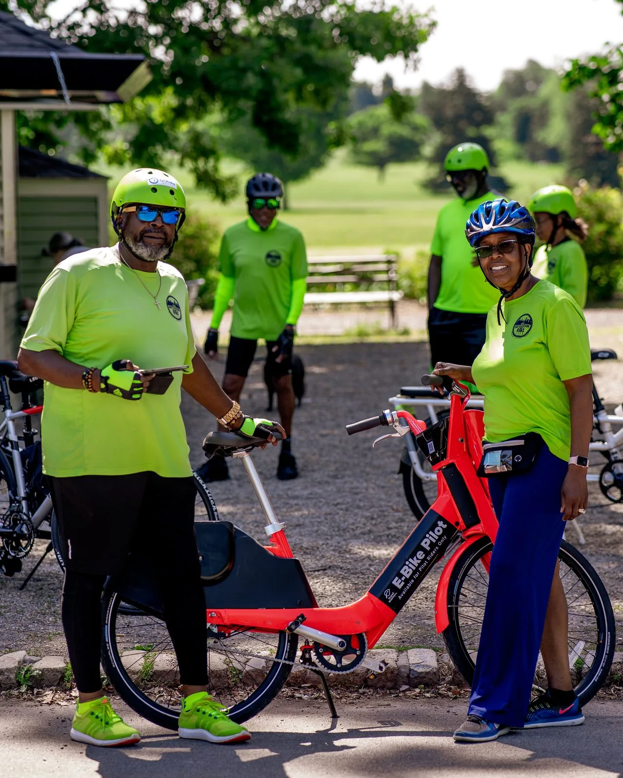 Two riders in high-visibility Shared Mobility shirts standing with a red E-Bike Pilot bike on a park path, with more riders and bicycles on the lawn behind them
