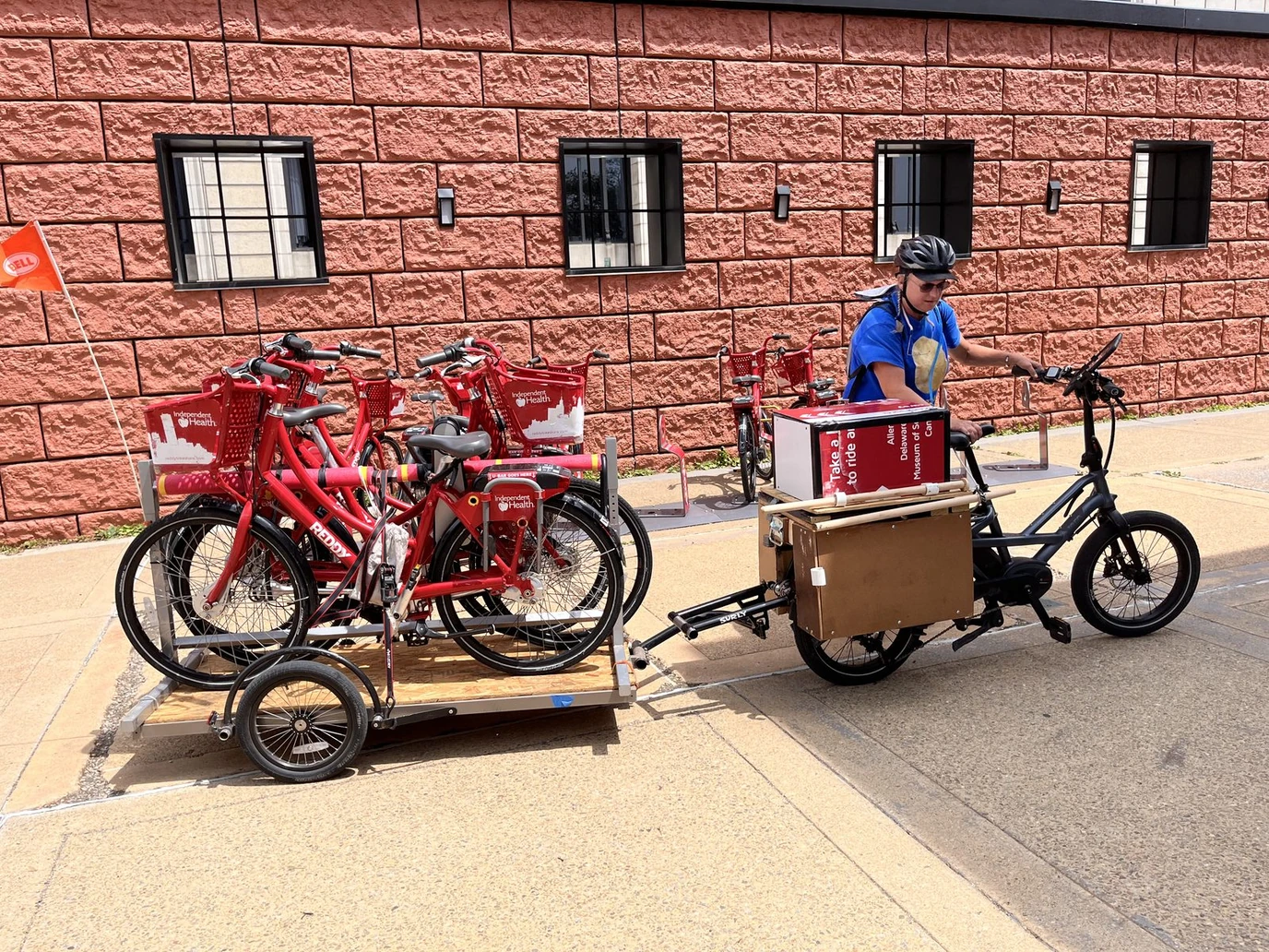 A crew unloading red bikeshare bicycles from a trailer beside a cargo e-bike