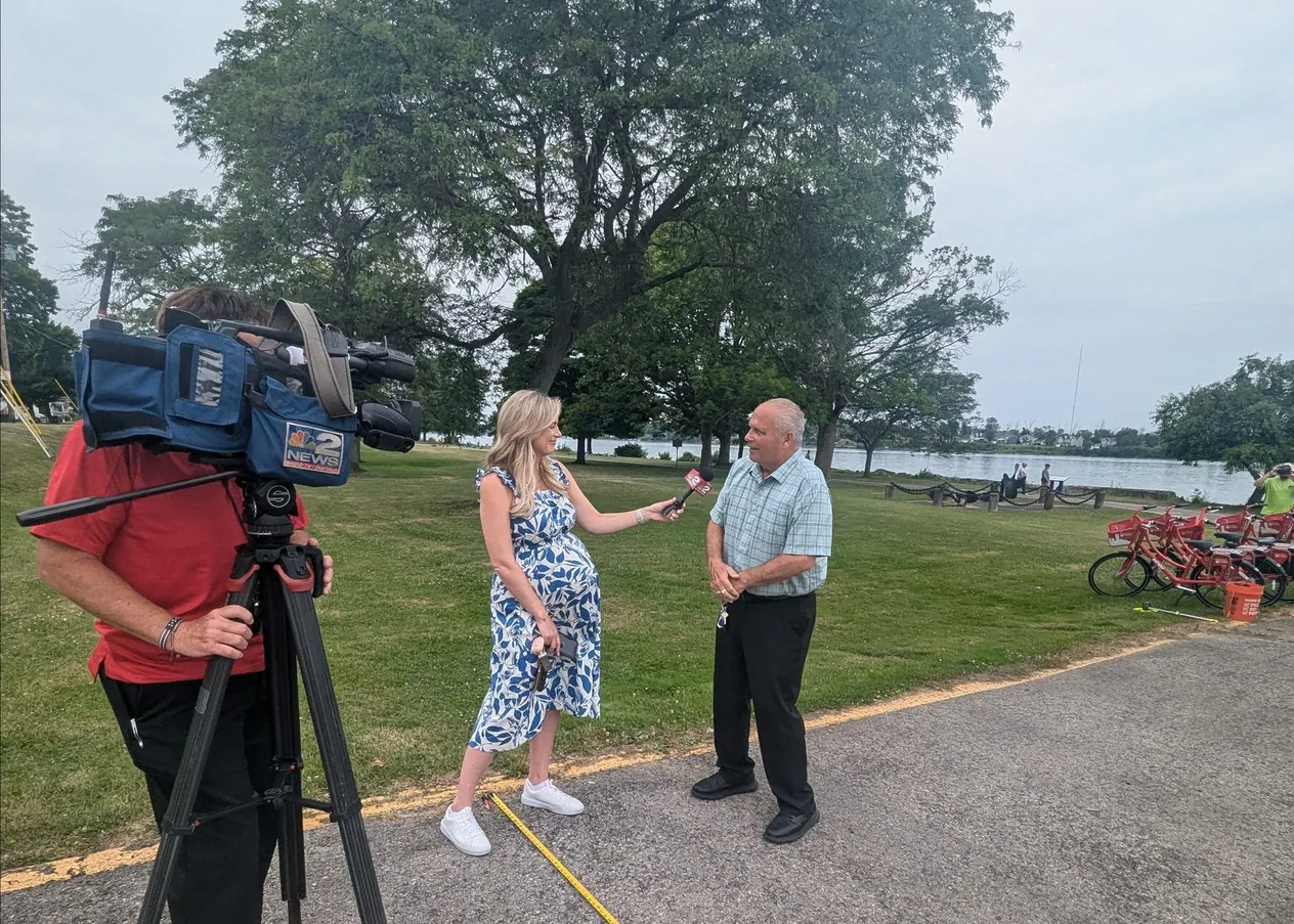 A television crew interviewing a man in a waterfront park, with Reddy bikes docked behind