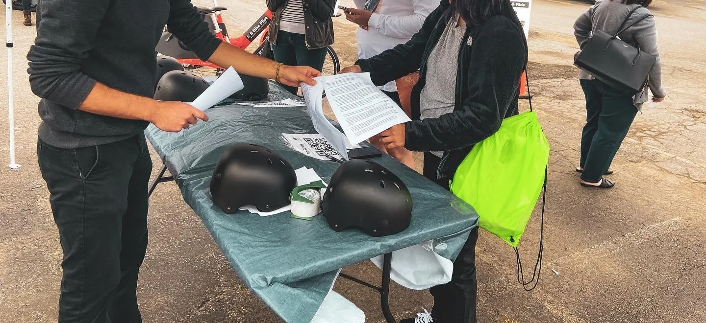Two people exchange paperwork across a table of black bike helmets at a Shared Mobility outreach event