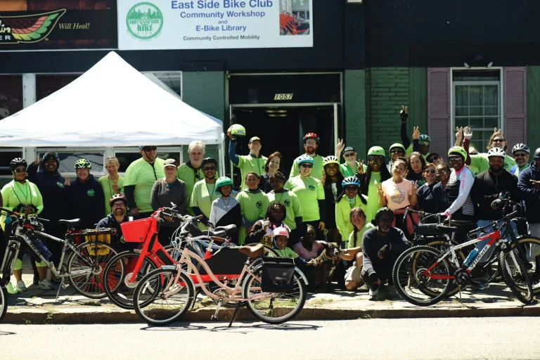 A large group of riders with bicycles outside the East Side Bike Club community workshop and e-bike library, under a Community Controlled Mobility banner