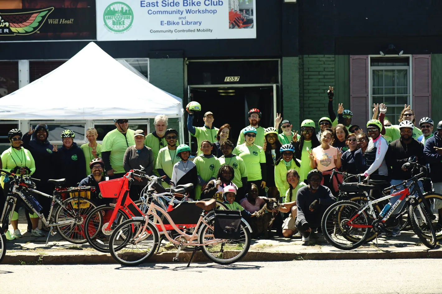 A large group of riders with bicycles outside the East Side Bike Club community workshop and e-bike library, under a Community Controlled Mobility banner