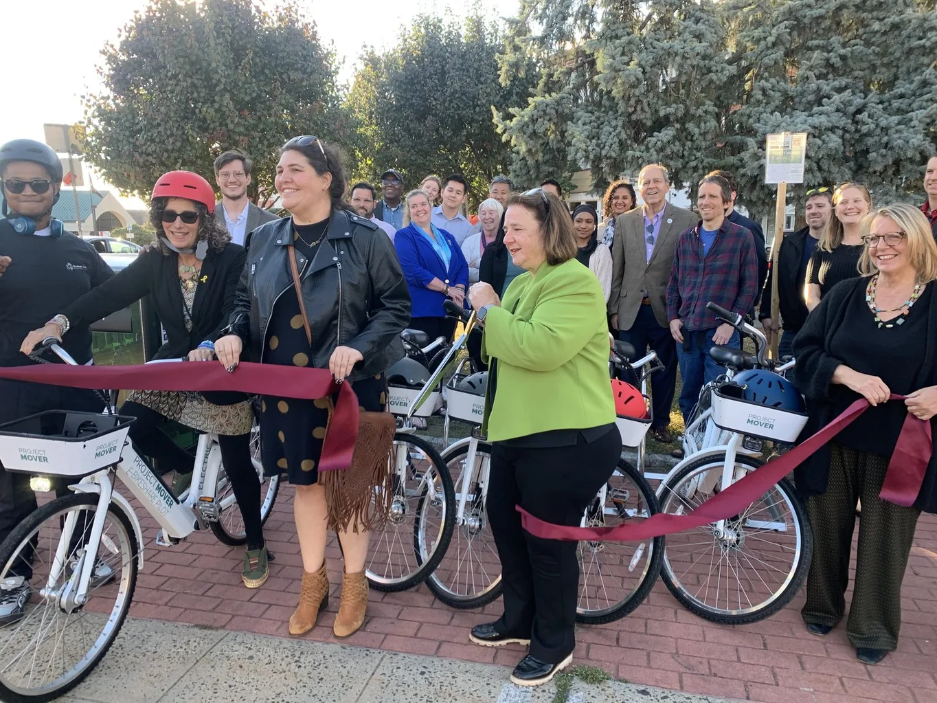 Officials cutting a ribbon in front of a row of white e-bikes at a program launch, with staff and partners gathered behind