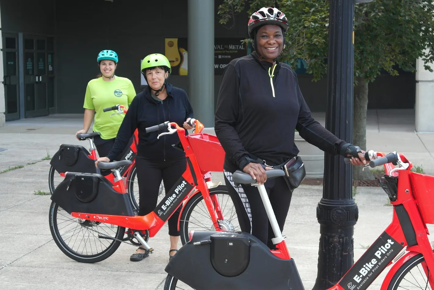 Three riders standing with red pilot e-bikes on a city street