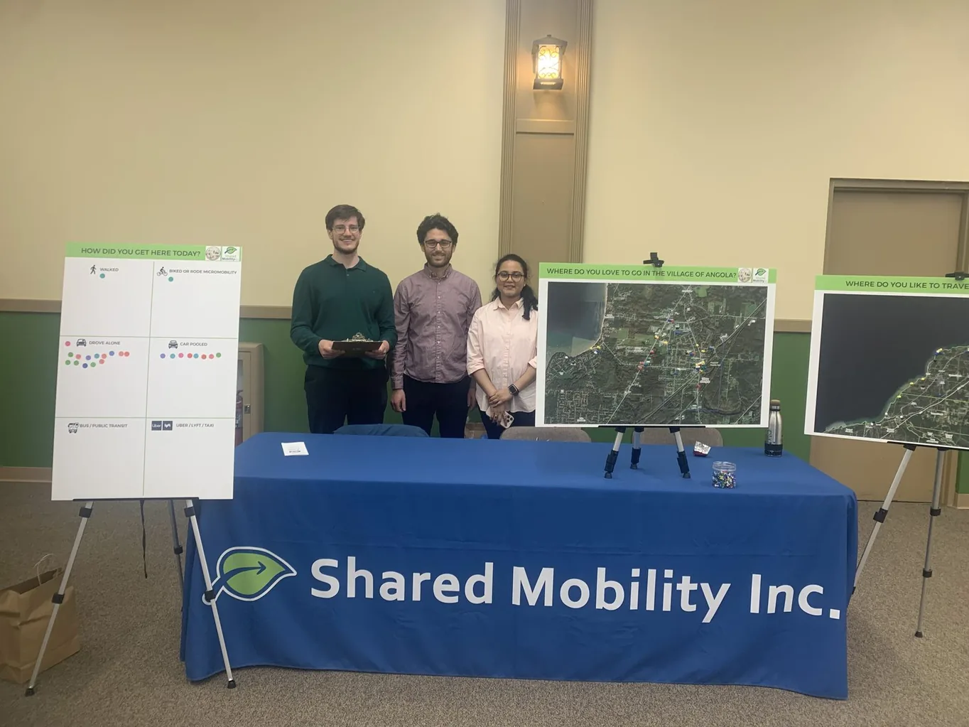 Three Shared Mobility Inc. staff behind a display table at a public engagement event, with travel-survey boards and maps on easels
